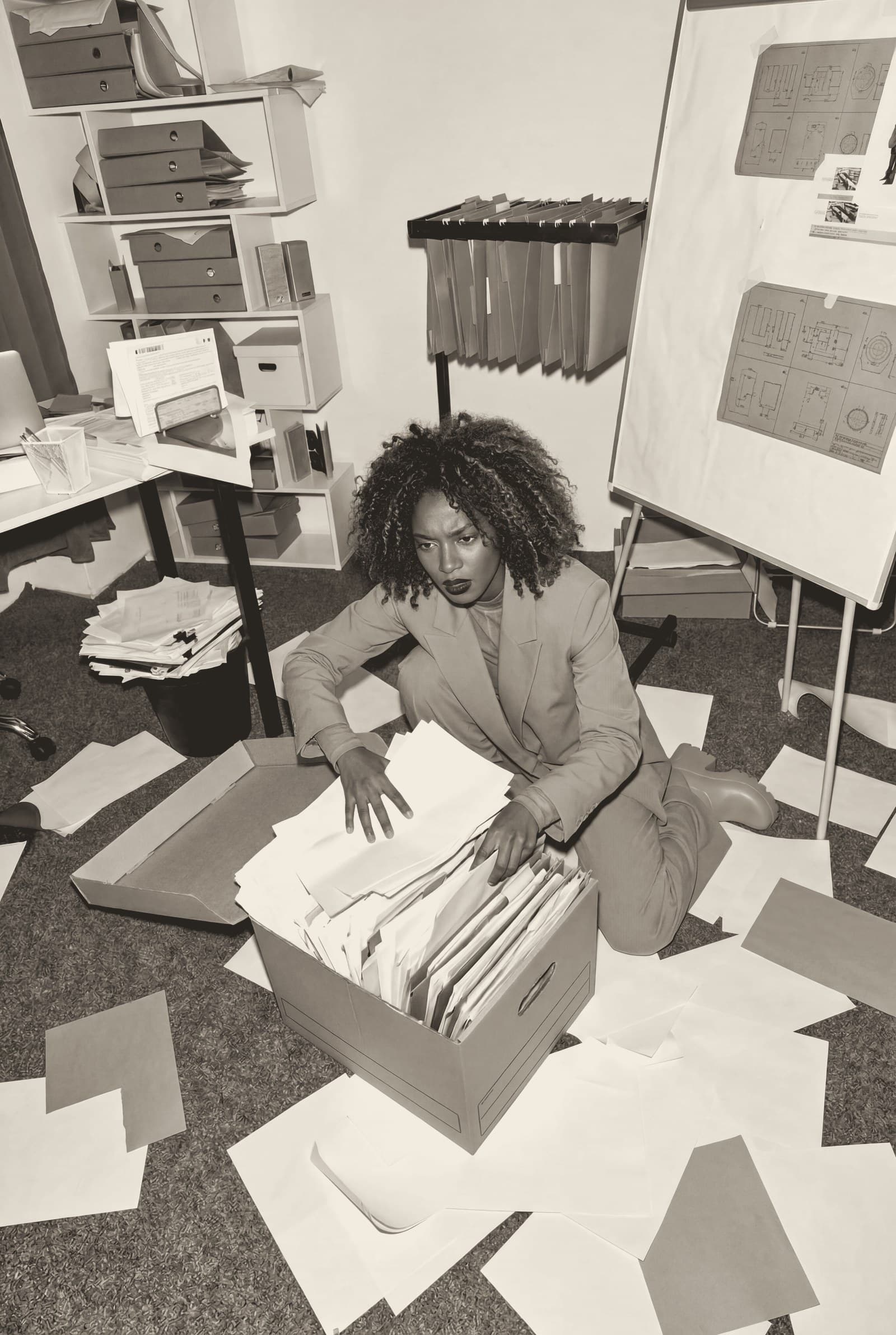 A woman seated on the floor of a cluttered office, sorting through a box of paperwork with papers scattered around her